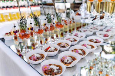 Çeşitli aperatifler ve tatlılarla dolu bir masa. Üstünde pembe çiçekler olan mini tartlar da dahil.