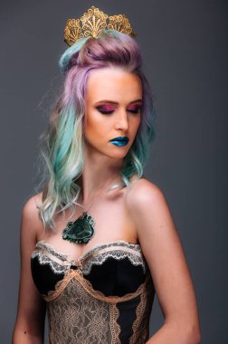 Mor saçlı, mavi dudaklı bir kadın taç ve kolye takıyor. Gri bir arka planın önünde duruyor.