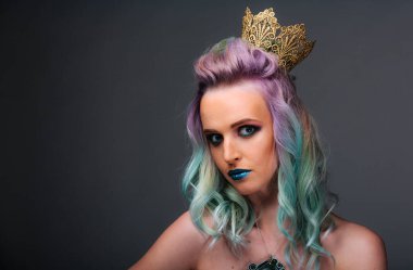 Mor saçlı ve kafasında altın taç olan bir kadın. Kameraya bakıyor.