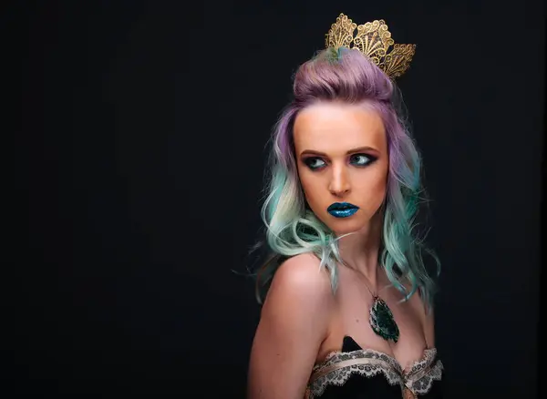 A woman with purple hair and blue lips stands in front of a black background. She is wearing a crown and a necklace