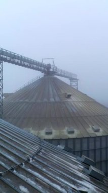 Sisli havada asansör kaplarının kirli çatıları. Tarım fabrikasının metal tankları arasında yüksekten uçuyor. İnsansız hava aracı görüntüleri. Dikey video