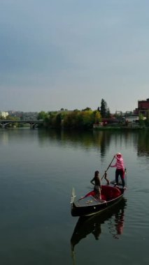 Gün batımında suda Gondol. Şehrin arka planında nehir boyunca yüzen bir kadın. Gondol kostümlü bir gondolcu. Dikey video