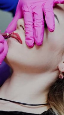 Güzellik salonunda dudak estetiği. Pembe lateks eldivenli usta müşterinin dudaklarını kırmızı kalemle çizer. Dudaklarda kalıcı makyaj. Dikey video