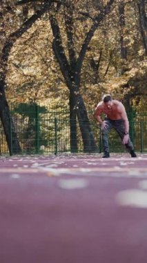 Stadyumda sağlıklı bir atlet koşuyor. Genç sporcu, sonbahar ağaçlarının arka planında koşuya katılıyor. Aşağıdan bak. Dikey video