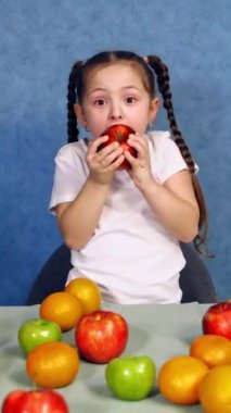 Lezzetli elma yiyen güzel küçük kız. Masada taze meyveler var ve komik bir çocuk mavi arka planda sulu kırmızı elmayı ısırıyor. Dikey video