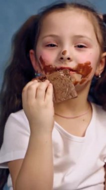 Kirli suratlı küçük kız çikolata yiyor. İki at kuyruklu komik çocuk mavi stüdyo arka planında lezzetli siyah çikolataları ısırıyor. Dikey video
