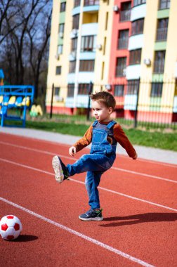 Genç bir çocuk bir pistte futbol topunu tekmeliyor. Çocuk mavi bir tulum ve kahverengi bir gömlek giyiyor.