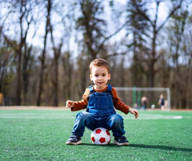 Genç bir çocuk önünde bir futbol topuyla çimlerin üzerinde oturuyor. Gülümsüyor ve eğleniyor.