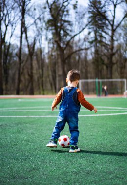 Mavi tulumlu genç bir çocuk bir sahada futbol oynuyor. Elinde kırmızı beyaz bir futbol topu tutuyor.