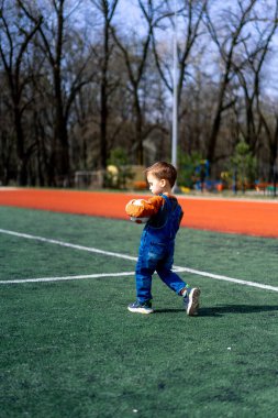 Genç bir çocuk sahada futbol topu tutuyor. Çocuk mavi bir elbise giyiyor ve eğleniyor.