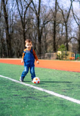 Genç bir çocuk sahada futbol oynuyor. Mavi gömlek ve kot pantolon giyiyor.