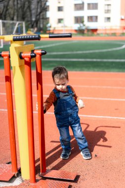 Genç bir çocuk sarı ve kırmızı bir fitness makinesinin önünde duruyor. Mavi tulum ve kahverengi gömlek giyiyor.