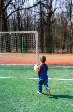 Genç bir çocuk sahada bir futbol topu tutuyor. Çocuk mavi bir tulum giyiyor ve yeşil bir tarlada duruyor.