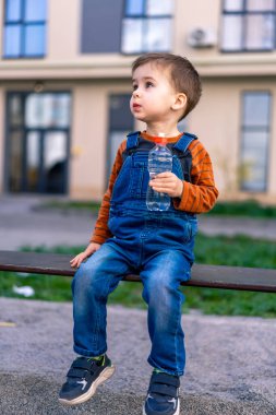 Genç bir çocuk elinde bir şişe suyla bankta oturuyor. Gökyüzüne bakıyor, muhtemelen bir şey düşünüyor.