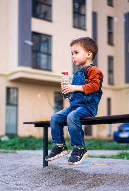 Genç bir çocuk elinde bir şişe suyla bankta oturuyor. Sahne, yakınlarda park etmiş bir arabayla bir parkta geçiyor..