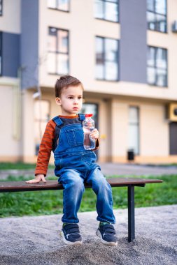 Genç bir çocuk bir bankta oturup su içiyor. Çocuk mavi bir tulum ve çizgili bir gömlek giyiyor..