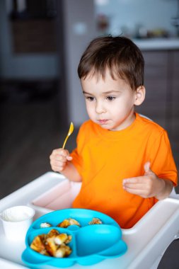 Genç bir çocuk mavi bir tabaktan yemek yiyor. Turuncu bir gömlek giyiyor ve elinde bir kaşık var.