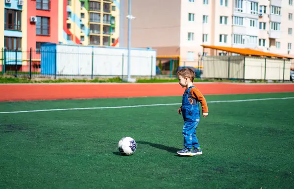 Genç bir çocuk sahada futbol topuyla oynuyor. Çocuk mavi bir tulum giyiyor ve çimlerin üzerinde duruyor.