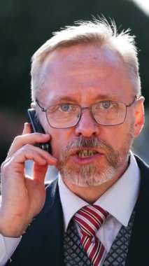 Gözlük takan ciddi bir işadamı kameraya bakarken telefonla konuşuyor. Şehirde takım elbiseli ve kravatlı bir adamın cep telefonuyla konuşurken portresi. Dikey video