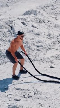 Atletik erkek, beyaz kayalık arka planda savaş halatları egzersizi yapıyor. Açık hava fitness antrenmanı sırasında şortlu sporcu kabloyla dalga yapıyor. Dikey video