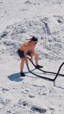 Sporcu antrenmanını dışarıda halatlarla bitirdiği için mutlu. Üstsüz sporcu, kayalık zeminde iki iple egzersiz yapıyor. Dikey video