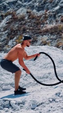 Vücut geliştirme eğitiminin yan görüntüsü, dışarıda dövüş halatları. Yakışıklı sporcu ellerini hızla dağ zemininde kabloyla hareket ettiriyor. Dikey video