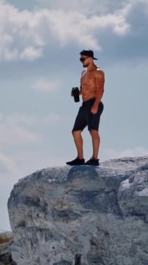 Şortlu kaslı bir erkek sadece dışarıda sıkı çalıştıktan sonra su içer. Beyaz kayanın üzerinde duran vücut geliştirici gökyüzü arka planında matarayla. Dikey video