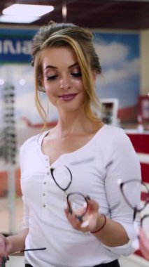 Güzel bir kadın aynanın önünde yeni gözlük deniyor. Sarışın bayan optik mağazada şık gözlükleri seçiyor ve mutlu bir şekilde gülümsüyor. Dikey video