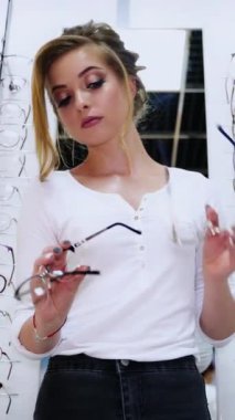 Güzel gülümseyen kız optik mağazada gözlük seçiyor. Güzel bir kadın arka planda yeni gözlükleriyle şık gözlük takmaya çalışıyor. Oftalmoloji. Dikey video