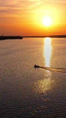 Gün batımında yüzen su ve motorlu tekne üzerinde hava manzarası. Suya yansıyan turuncu güneşin batışını görmek çok güzel. Nehirdeki tekne sanayi fabrikasına gidiyor. Dikey video