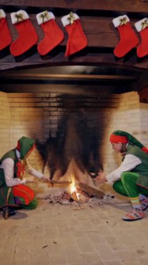 Noel arkaplanı, oturma odasındaki şöminenin yanında oturan iki elf. Şöminenin üzerinde kırmızı çoraplar asılı ve periler ellerini içeride ateşe yakın ısıtıyorlar. Dikey video