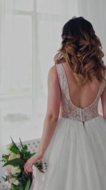 Işık stüdyosunda güzel bir modelin üzerindeki seksi dantelli gelinlik. Lüks elbiseli, elinde bir buket çiçekle çekici gelin yatak odasının penceresine geliyor. Dikey video