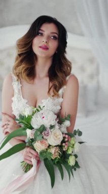 Seksi gelinlikli çekici genç gelin, yatakta oturuyor. Çiçekli güzel gelin. Beyaz elbiseli güzel manken yatak odasındaki kadın fotoğrafçıya poz veriyor. Dikey video