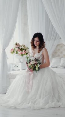 Beyaz dantel elbiseli güzel genç manken beyaz bir yatakta oturuyor. Çiçekli gelinlik içindeki muhteşem gelin aydınlık yatak odasında oturuyor. Dikey video