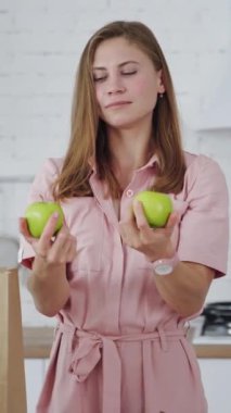Yeşil elma tutan açık renk elbiseli güzel bir kadın. Modern mutfakta taze meyveli genç ve mutlu bir kadın kameraya bakıyor. Sağlıklı beslenme. Dikey video