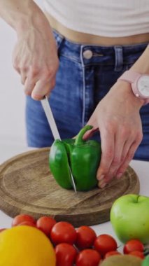 Genç bir kadın ahşap bir tahtada yeşil biber kesiyor. Yakın plan kadın elleri mutfakta taze malzemeler üzerine sağlıklı yiyecekler hazırlıyor. Dikey video