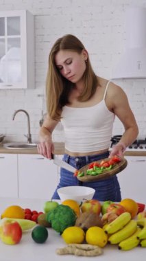 Genç ve güzel bir kadın modern mutfakta lezzetli salata hazırlıyor. İçerideki masada bir sürü taze organik malzeme var. Sağlıklı yiyecekler. Diyet konsepti. Dikey video