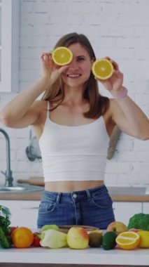 Mutfakta portakallı genç bir kadın ve masada bir sürü taze meyve ve sebze. İki yarım portakal tutan güzel dişi gözlerini kapatıp neşeyle kameraya bakıyor. Dikey video
