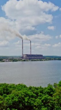 Yukarıdan nehir manzarası ve iki fabrika borusu. Yazın bir nehrin kıyısındaki elektrik santrali borularından duman çıkıyor. Hava görüntüsü. Dikey video