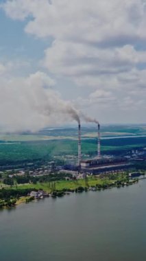 Yazın nehrin kıyısındaki bitkinin havadan görünüşü. Yukarıdan güzel nehrin doğal arka planına ve borulardan duman çıkan fabrikaya bakın. Ağır çekim. Kamera yukarı çıkıyor. Dikey video