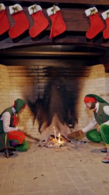 Noel arka planında şöminenin yanındaki elfler içeride. Yeşil kostümlü komik elfler ateşin yanında oturuyorlar. Dikey video