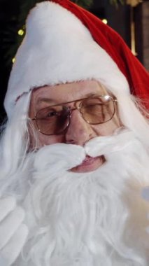 Noel Baba 'nın gözlüklü portresi kameraya bakıyor. Yaşlı Noel Baba, beyaz sakallı ve gözlükleriyle gözlerini kırpıyor. Yakın plan. Dikey video