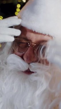 Beyaz sakallı, gözlüklü, yakın plan Noel Baba. Noel Baba 'nın beyaz ve kırmızı kostümlü portresi evin içinde. Dikey video