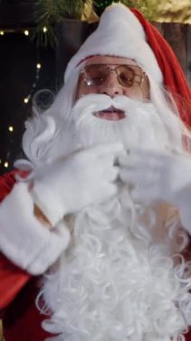 Uzun beyaz sakallı Noel Baba şarkı söylüyor. Noel Baba kırmızı kostüm ve beyaz eldivenler giyip Noel arka planında sakalını düzeltiyor. Dikey video