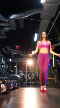 Pembe bluzlu, taytlı spor bir kadının fitness merkezinde ip üstünde zıplaması. Modern simülatörlerin arka planında ip atlama eğitimi almış sıska bir genç kız. Dikey video