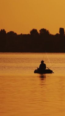 Gün batımında bir balıkçıyla tekne gölde yavaşça yüzüyor. Teknede oturan ve nehirde güzel bir gün batımının turuncu arka planında balık tutan bir adamın silueti. Dikey video