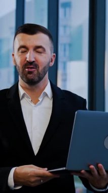 Gülümseyen sakallı, takım elbiseli iş adamı elinde dizüstü bilgisayarla pencerenin yanında duruyor. Adam elleriyle bir şey göstererek konuşuyor. Dikey video