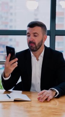 Masada oturan erkek işadamı bir telefon görüşmesi yapacak. Adam not almak için kağıt bir defter ve kalem alır. Ofis arkaplanı. Dikey video