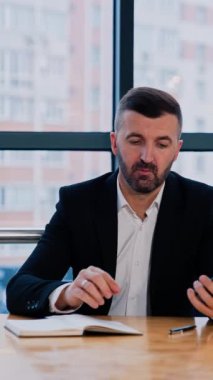 Takım elbiseli sakallı adam ofisindeki masada oturuyor. İş adamı telefonuna bakıyor, bilgi topluyor. Bulanık şehir arkaplanı. Dikey video