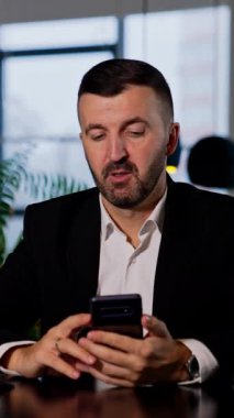 Erkek ofis çalışanı iyi bir ruh hali içinde telefonuna bakar. Adam telefonuna bir şey kaydırıyor ve bu süreçte yorum yapıyor. Arkaplan bulanık. Dikey video